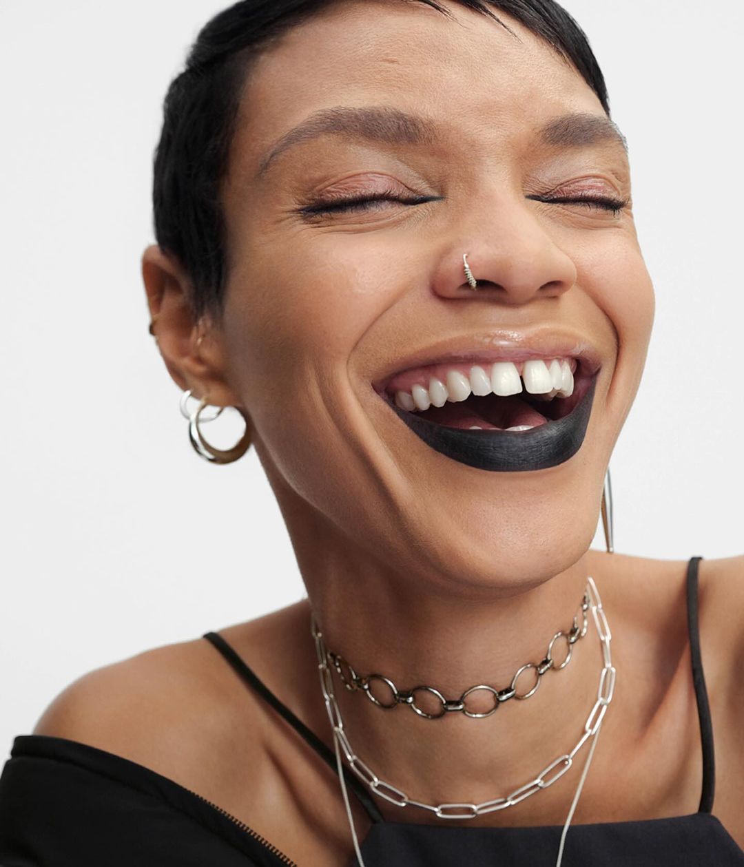 Close-up of a smiling adult woman with dark lipstick, silver necklaces, and hoop earrings against a bright white background.
