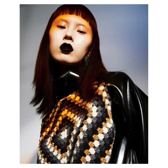 Striking studio shot of an Asian female model with dark lipstick, bowl cut bangs, wearing a black and patterned top against a graded grey background.
