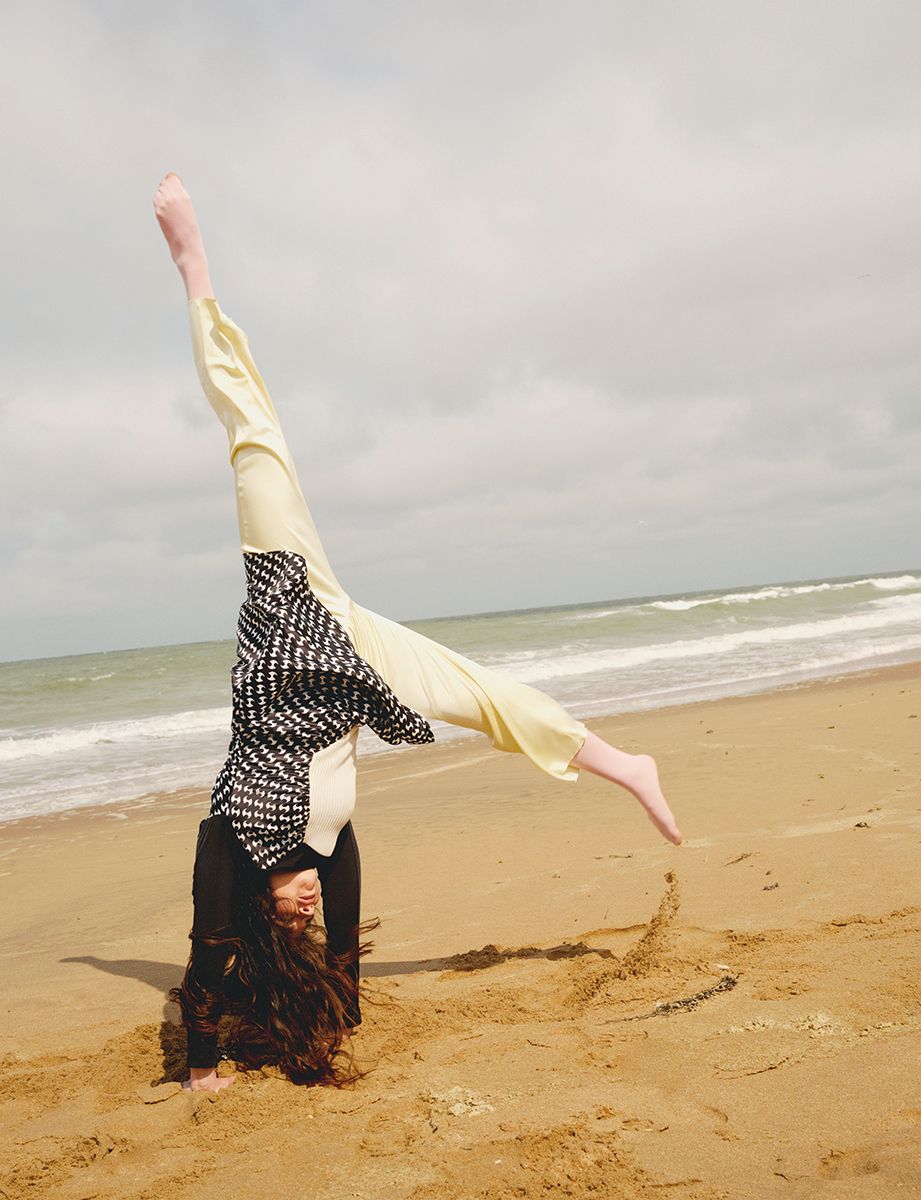 A woman performs a handstand on a sandy beach with ocean waves in the background, showing off her long hair and casual clothing.