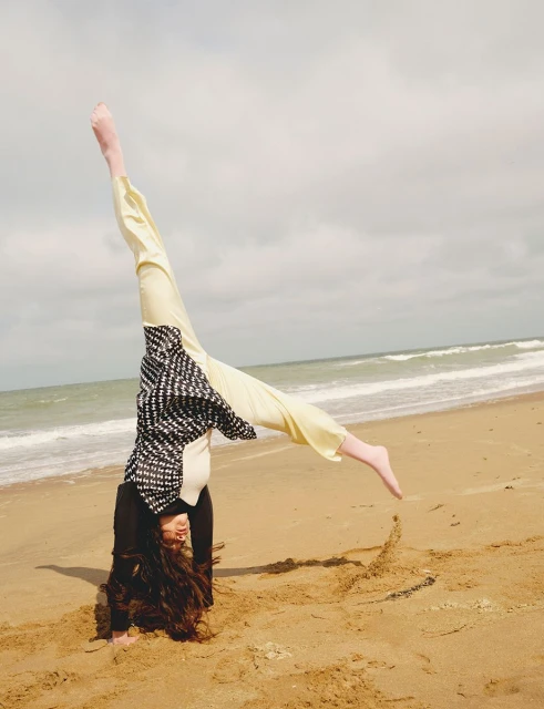 A woman performs a handstand on a sandy beach with ocean waves in the background, showing off her long hair and casual clothing.