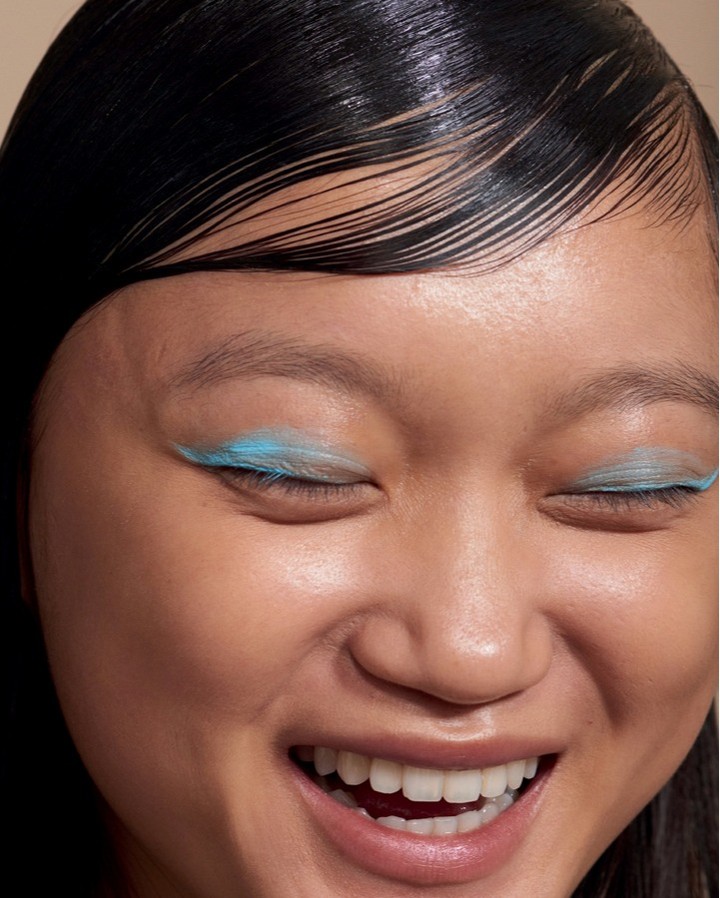 Close-up of a smiling young Asian woman with bold blue feather eyelashes and wet-look black hair, capturing a modern beauty aesthetic.
