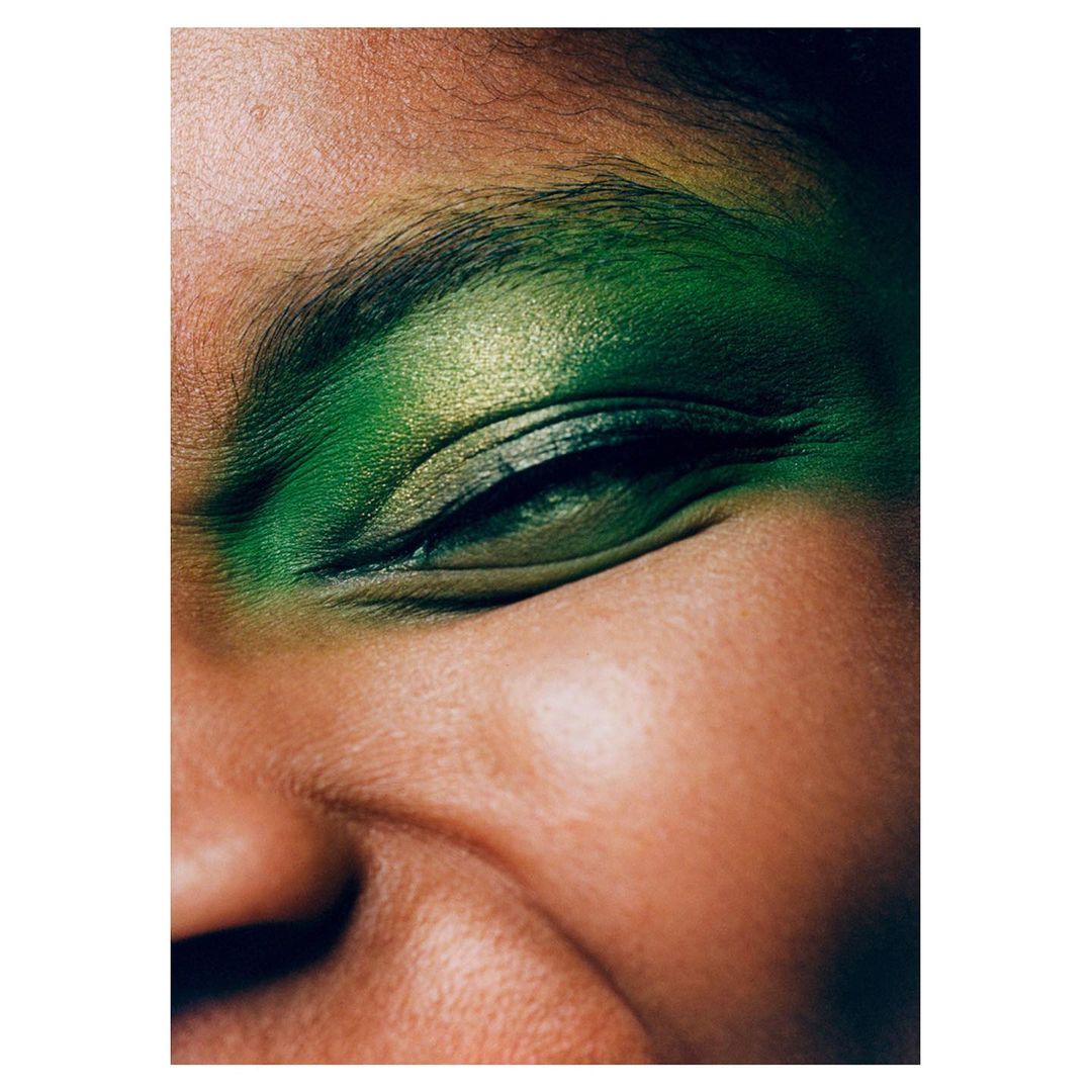 Close-up portrait of a woman's face featuring bright, green eye shadow and fair skin. The shot captures natural light and texture.