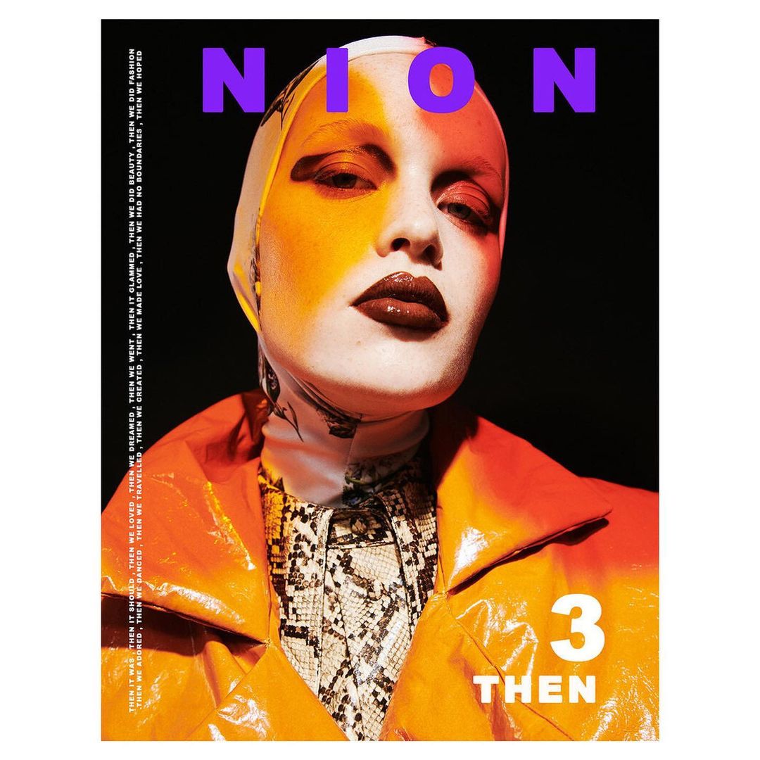 Fashion editorial shot of a woman in an orange coat and patterned scarf, featuring bold makeup and neon text overlay on a dark background.