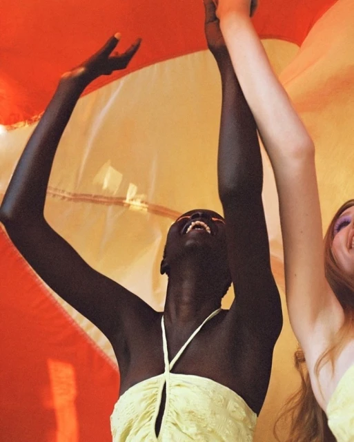 Two women laugh with arms raised against a backdrop of orange and yellow fabric, wearing light green tops, showcasing a joyful mood.