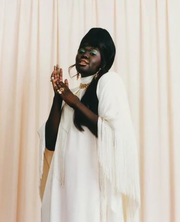 Black woman in a white fringed cape and turban rubbing hands together against draped fabric background