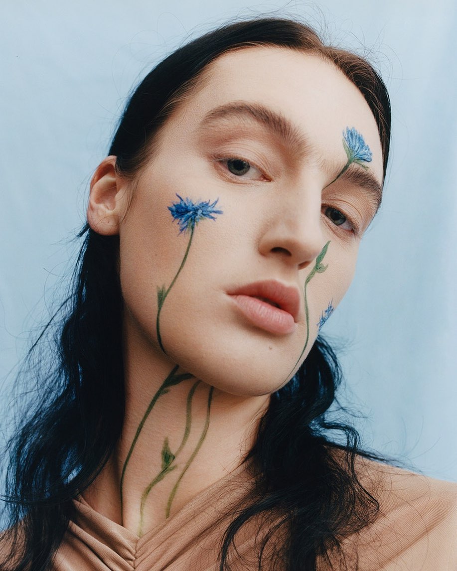 Close-up portrait of a female model with cornflower-inspired face paint on a light blue background in a beauty and fashion editorial style.