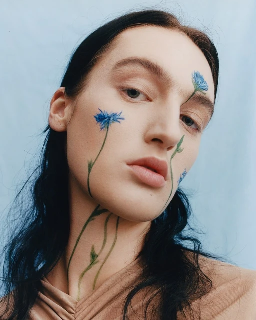 Close-up portrait of a female model with cornflower-inspired face paint on a light blue background in a beauty and fashion editorial style.