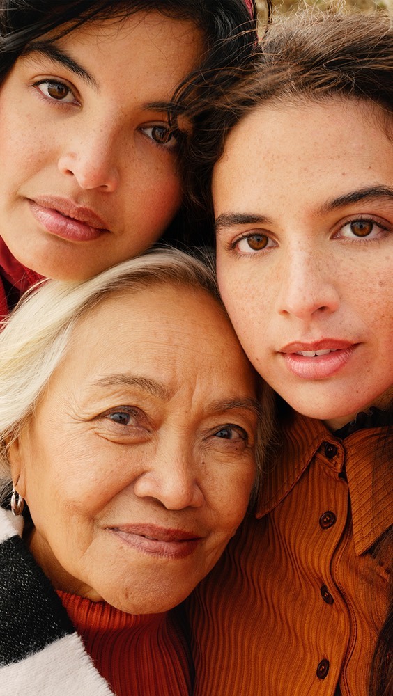 Close-up of three diverse women, one elderly, posing closely together outdoors in natural light, creating a warm, familial portrait.