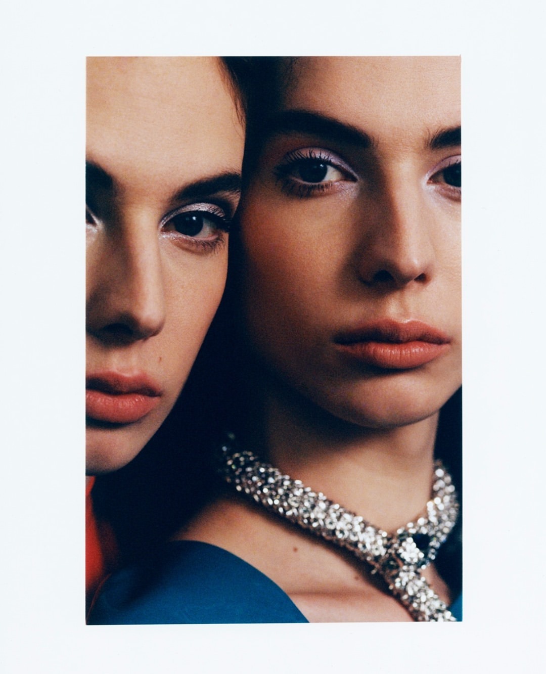 Close-up studio shot of two female fashion models with neutral expressions, wearing blue tops and rhinestone necklaces against a white backdrop.