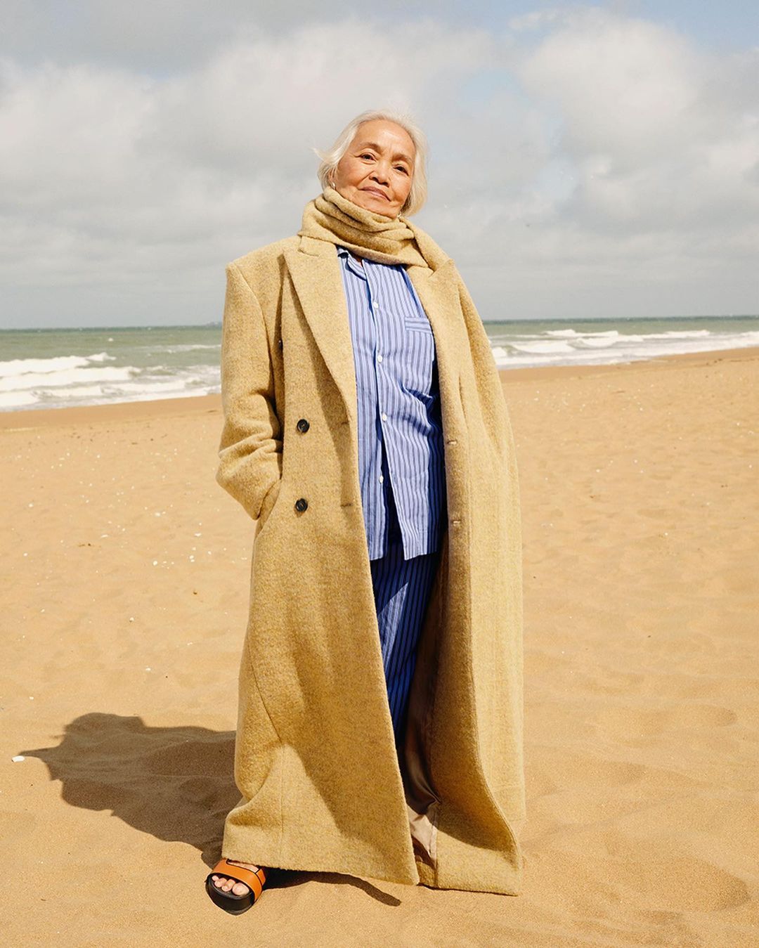 Elegant elderly woman in oversized tan coat and blue-striped pajamas on a sandy beach under a cloudy sky, looking at the camera.