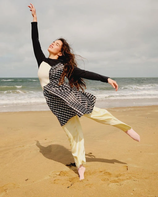 Adult female dances barefoot on the beach in a black and white top with long sleeves and light yellow pants near the ocean.