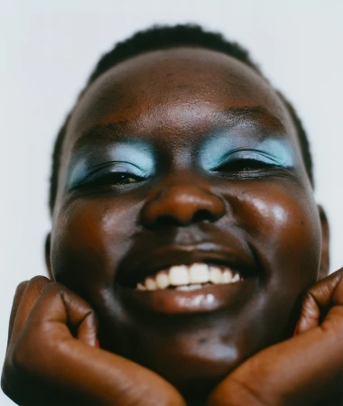 Close-up portrait of a smiling Black woman with blue eyeshadow resting chin on hands