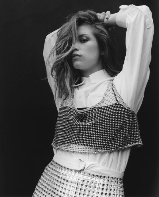 Black and white portrait of a blond woman in a white button-down, silver top, and skirt against a dark background, hands above head.