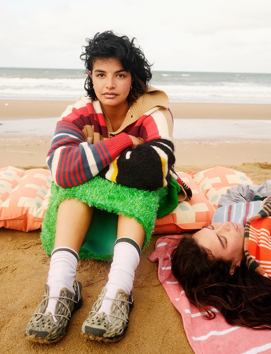 Two women pose on a beach with a pastel blanket and pillows, one sitting, one laying, wearing colorful sweaters and sporty shoes.