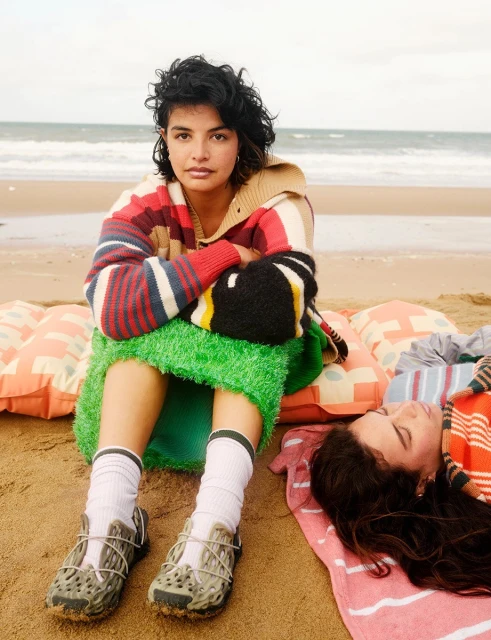 Two women pose on a beach with a pastel blanket and pillows, one sitting, one laying, wearing colorful sweaters and sporty shoes.
