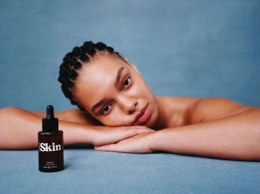Woman poses with skincare product against a blue backdrop in a beauty shot, highlighting the bottle and model's radiant skin.