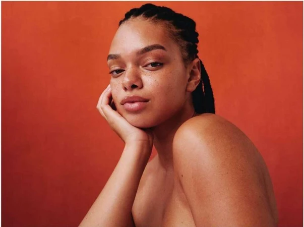Portrait of a young black woman with braided hair and freckles resting her head on her hand against an orange background.