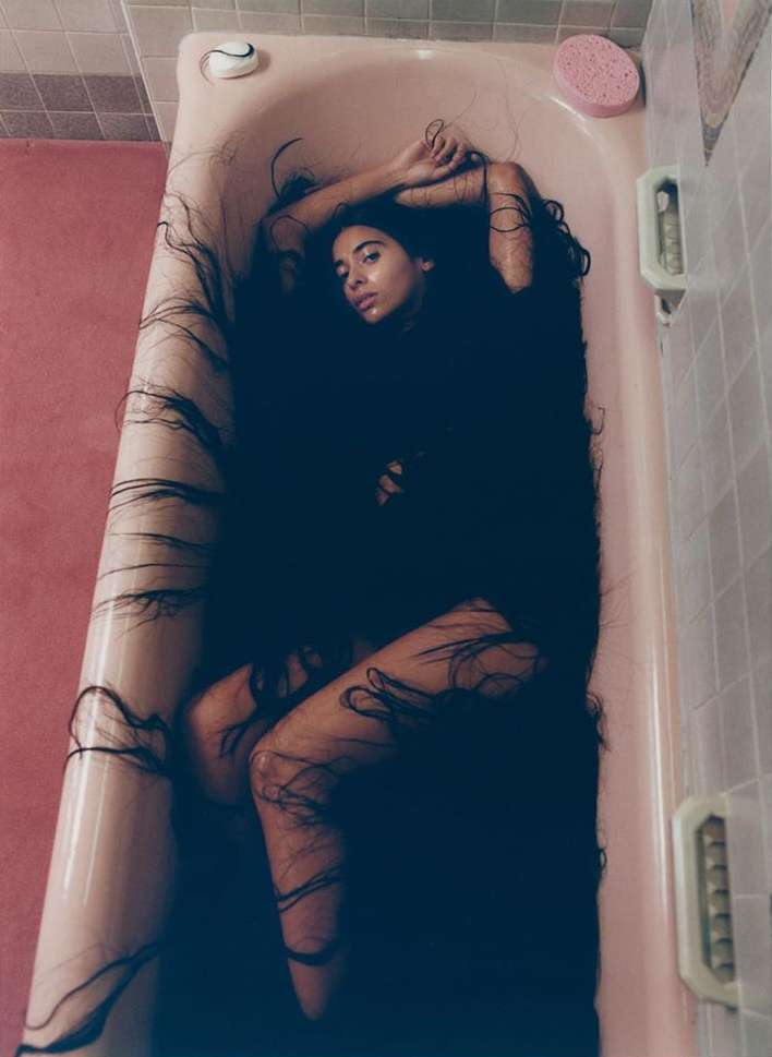 Woman with long dark hair reclines in a pink bathtub in a tiled bathroom, creating an artistic and unusual portrait.