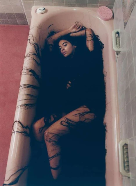 Woman with long dark hair reclines in a pink bathtub in a tiled bathroom, creating an artistic and unusual portrait.