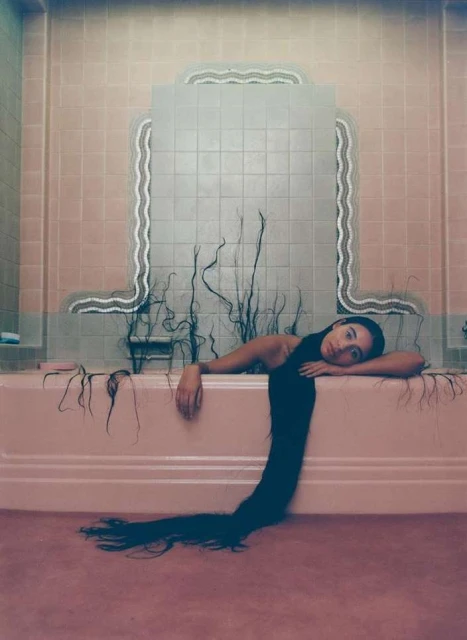 Woman reclines in a pink bathtub, her long black hair cascading over the edge onto the pink floor, set against a pink-tiled bathroom wall.