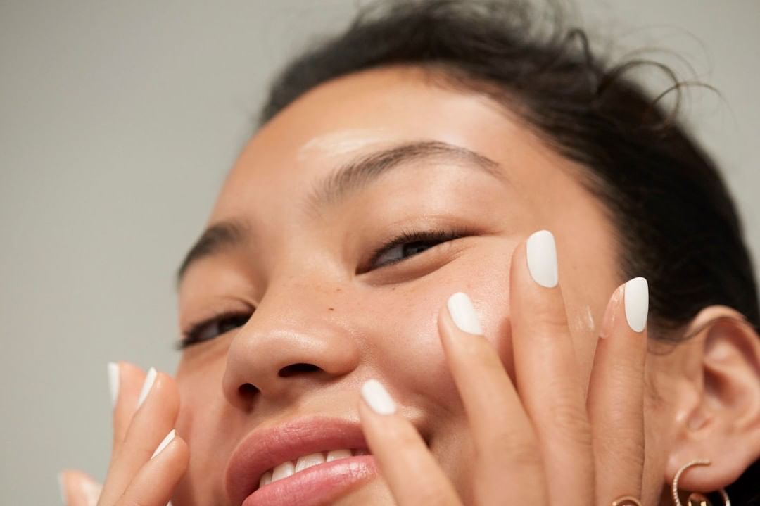 Close-up shot of an Asian woman smiling and applying skincare to her face. She has white nails and gold hoop earrings.