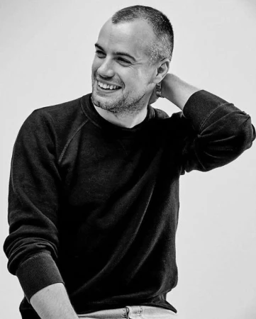 Monochrome portrait of a smiling man with short hair wearing a dark sweatshirt, hand touching the back of his neck.