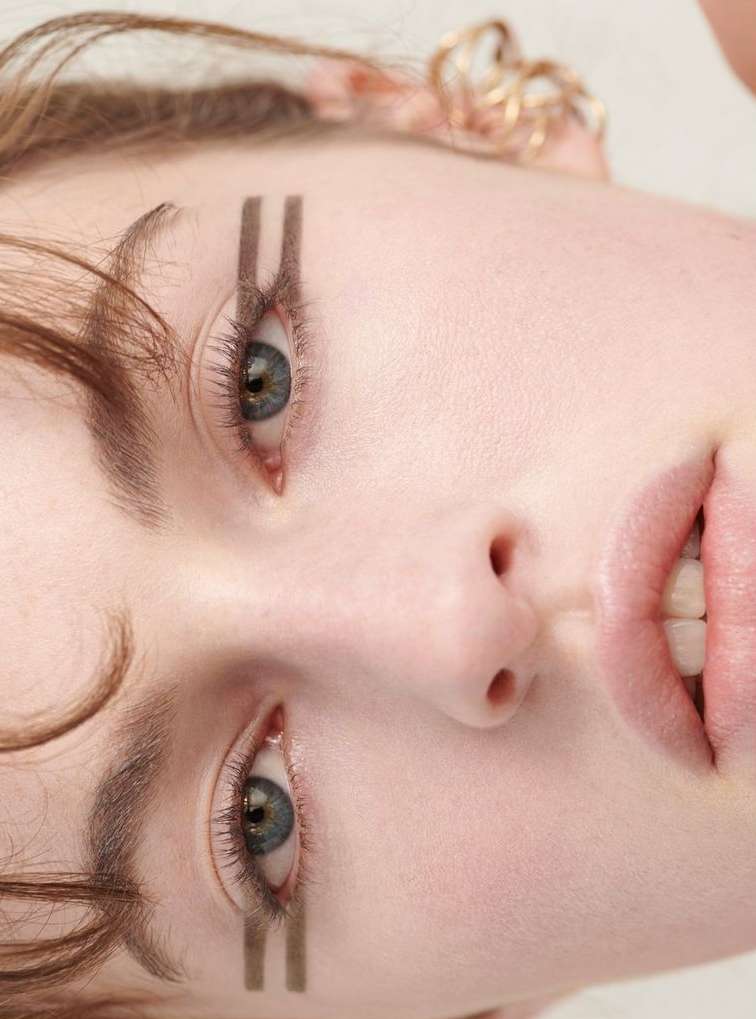 Close-up portrait of a fair-skinned young adult woman with green eyes, wearing brown eye makeup and gold jewelry in a photo studio.