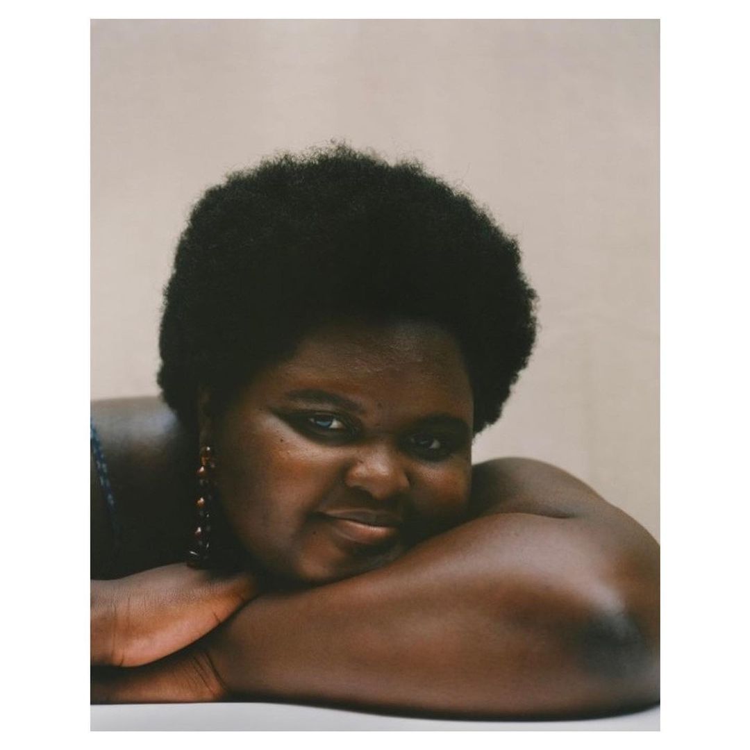 Portrait of a smiling African-American woman with dark afro, arms crossed, wearing earrings, and set against a neutral, soft pink background.