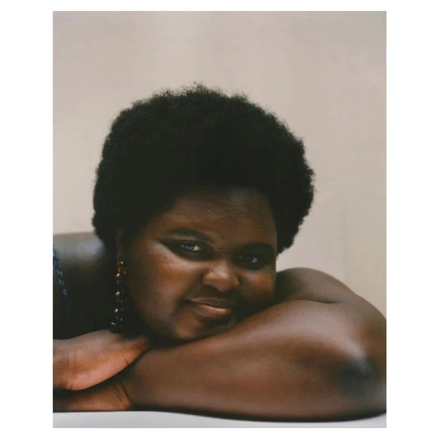 Portrait of a smiling African-American woman with dark afro, arms crossed, wearing earrings, and set against a neutral, soft pink background.