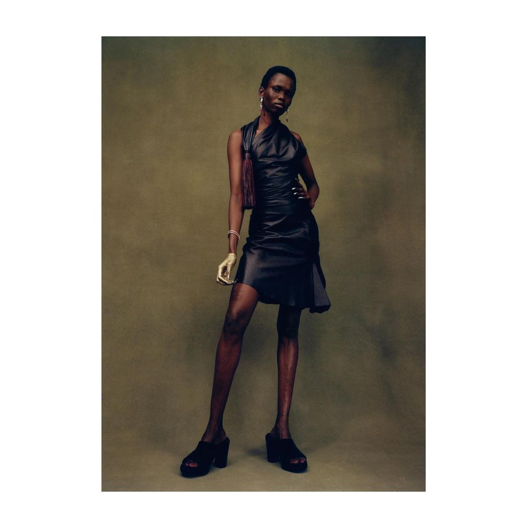 A stylish Black female model poses in a shiny dark dress with a side bag, wearing platform shoes against a textured olive background.