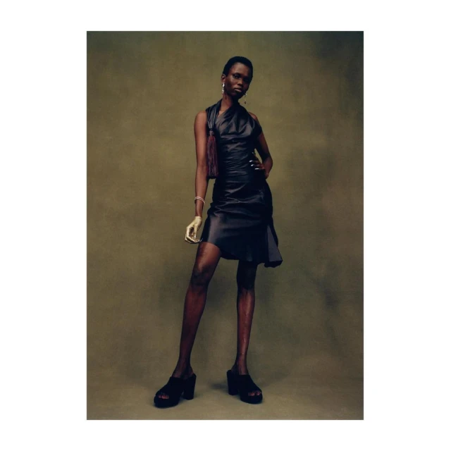 A stylish Black female model poses in a shiny dark dress with a side bag, wearing platform shoes against a textured olive background.