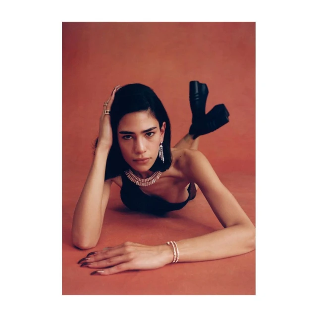 A woman with dark hair poses lying down wearing jewelry and black top on a coral colored background in a fashion photography style.