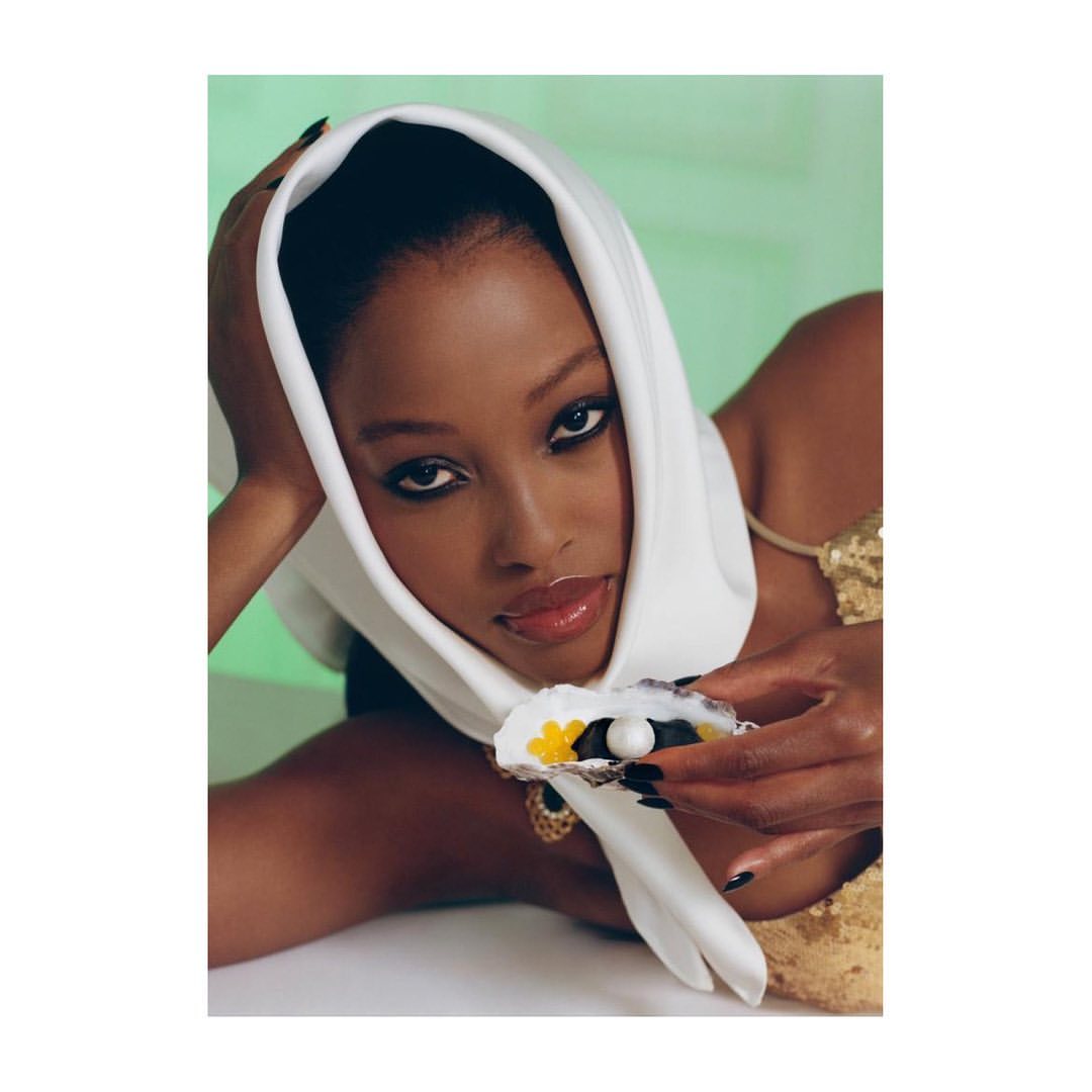 Elegant portrait of a black woman wearing a white headscarf, gold necklace, and holding an oyster shell with a pearl and caviar.