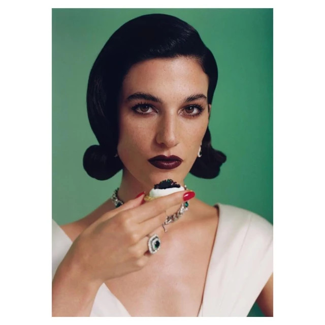 Elegant woman with dark hair poses against a green backdrop, adorned with emerald jewelry, holding a canape topped with black caviar.
