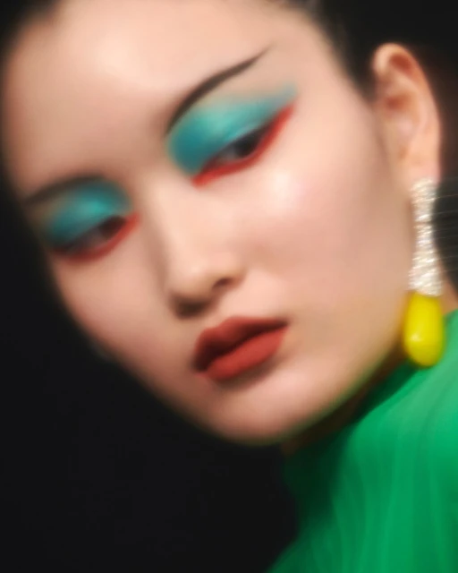 Close-up of a woman with vibrant blue and red eyeshadow, dramatic black eyeliner, glossy red lipstick, and a green top.