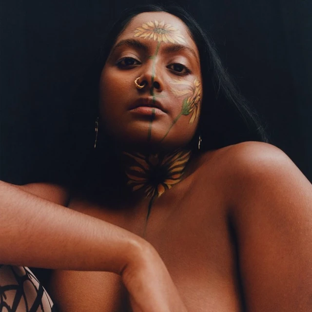 Woman with sunflower face paint gazes intently at the camera. Dark hair frames her face, and a black background enhances the portrait.