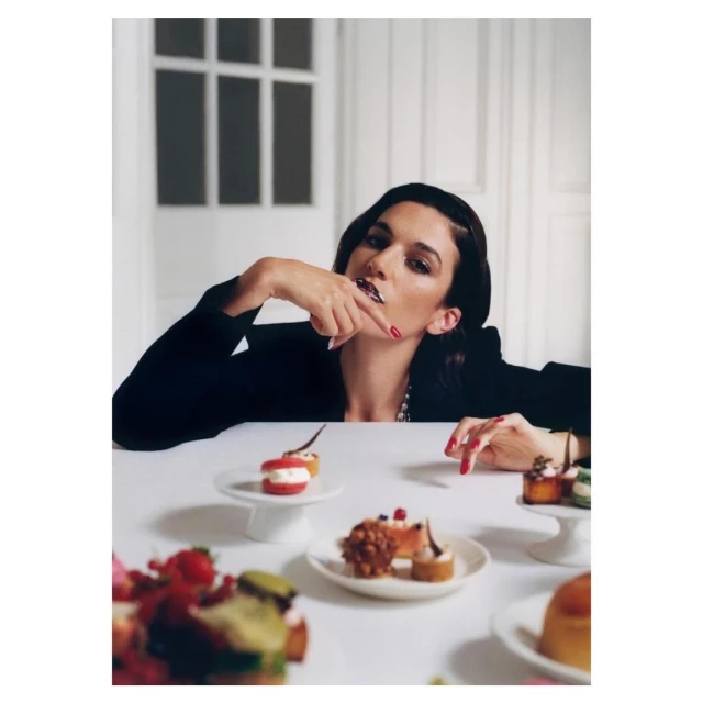 Woman in a black blazer sits at a table with assorted desserts on plates in a bright interior. She is holding her finger to her lip.