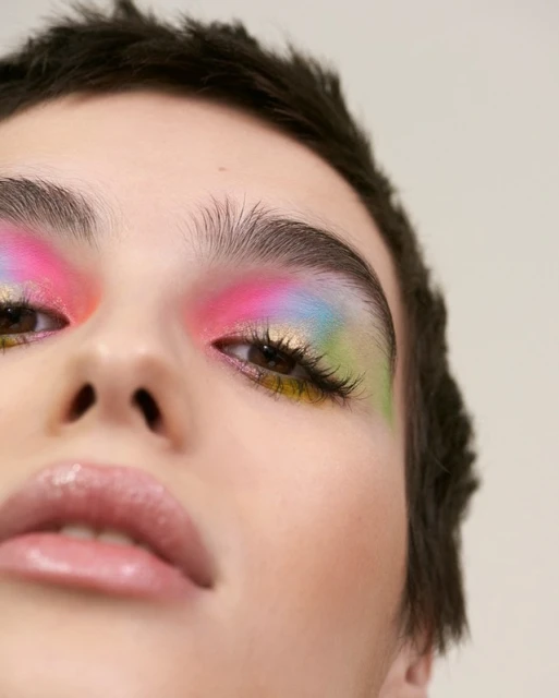 Close-up beauty shot of a woman wearing colourful eye makeup with pink, yellow, blue and green shades against a neutral background.