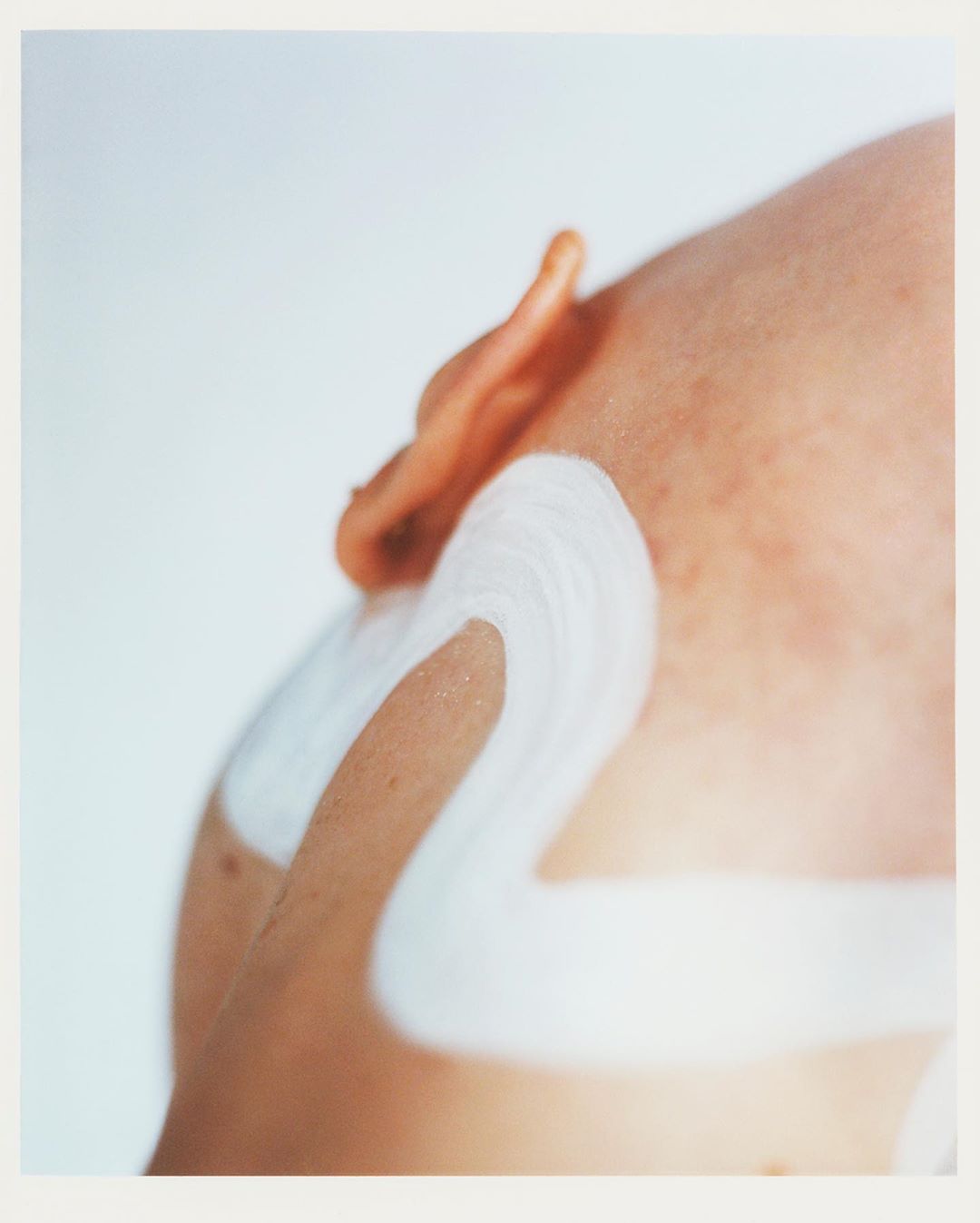 Close-up studio shot of a person's shoulder and ear with white paint or cream applied in broad strokes against a clean white backdrop.