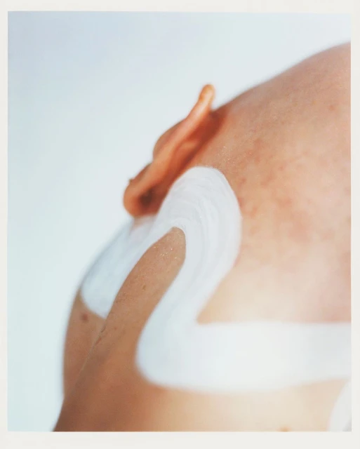 Close-up studio shot of a person's shoulder and ear with white paint or cream applied in broad strokes against a clean white backdrop.