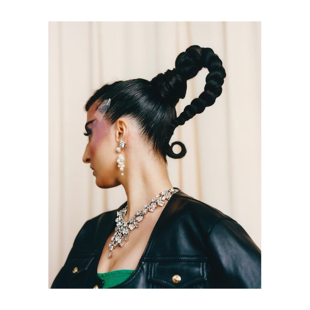 Profile shot of a woman with an elaborate braided hairstyle, wearing jewelry and a black leather jacket against a neutral backdrop.