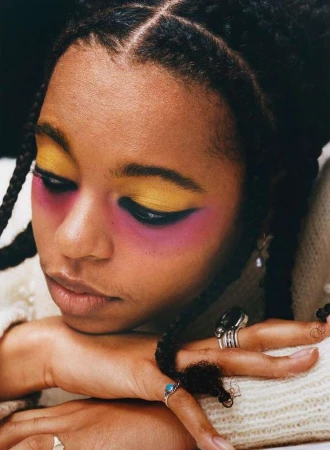 Close-up of a Black woman with yellow and pink eye makeup, braided hair, multiple rings, wearing a textured sweater.