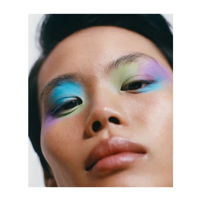 Close-up studio shot of an adult female with colorful pastel blue, green, and purple eyeshadow, fair skin and black hair.