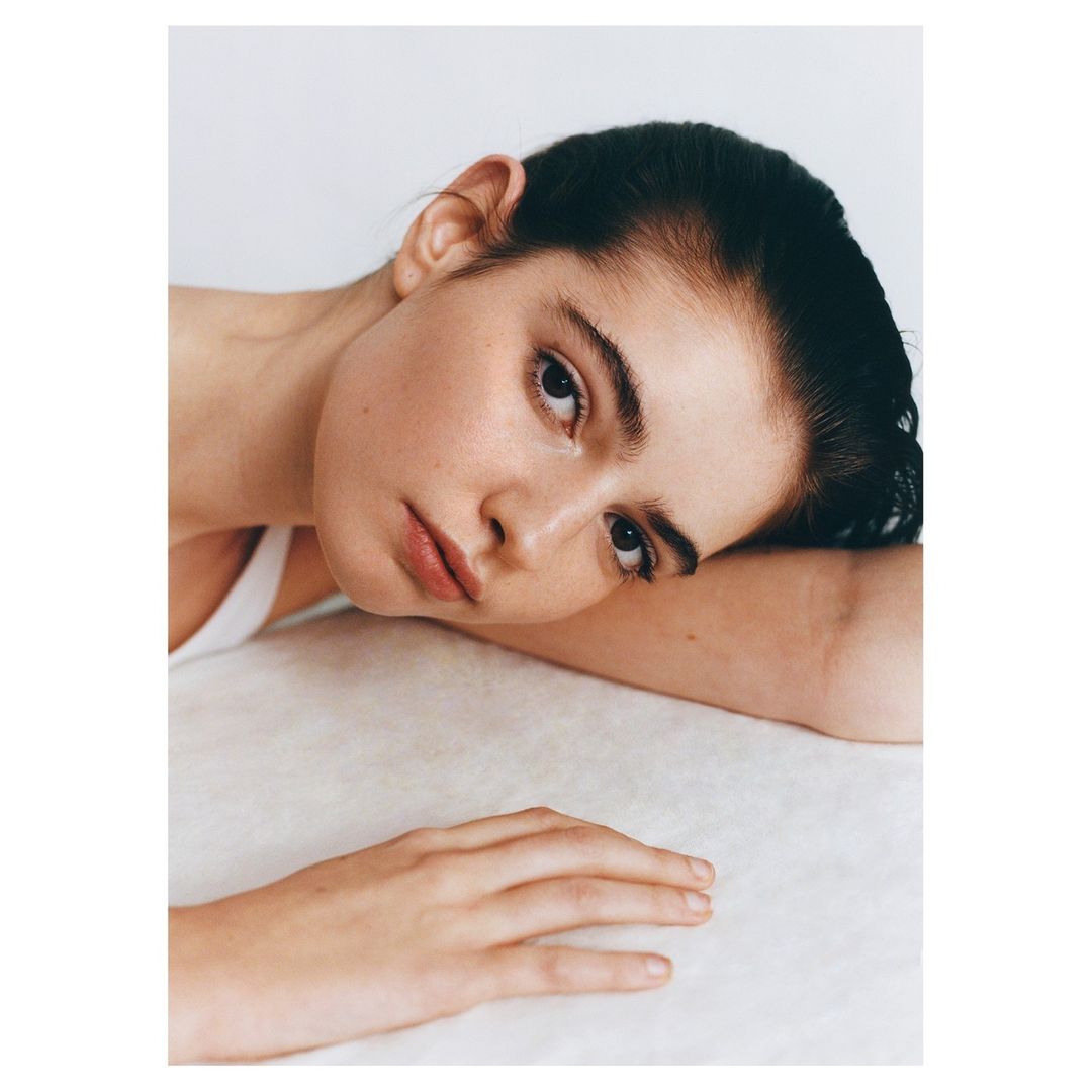 Close-up portrait of a woman with dark hair resting her head on her arm against a white background