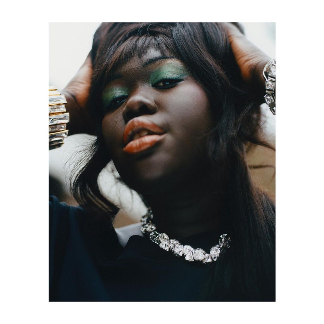 Close-up portrait of a woman with green eye shadow, black hair, and a silver necklace, striking a pose with her hands in her hair.