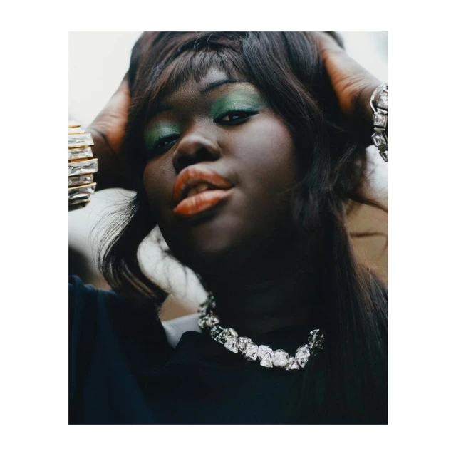 Close-up portrait of a woman with green eye shadow, black hair, and a silver necklace, striking a pose with her hands in her hair.