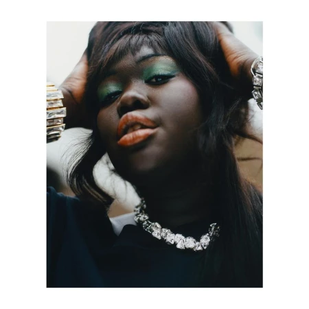 Close-up portrait of a woman with green eye shadow, black hair, and a silver necklace, striking a pose with her hands in her hair.