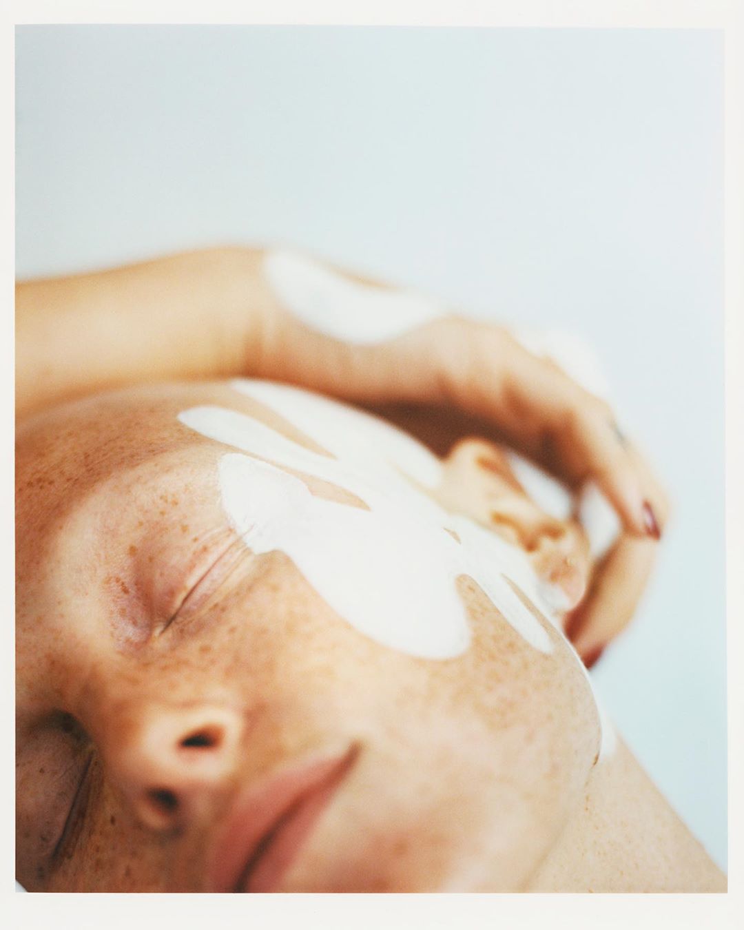 Close-up portrait of a freckled person with white cream or mask smeared on face and arm against a plain white background