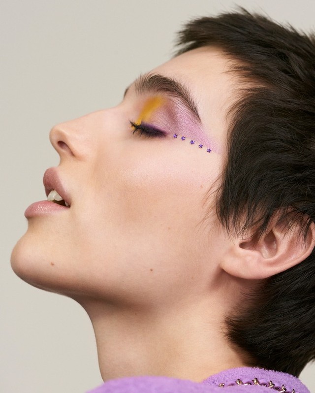 Close-up studio shot of a woman with creative yellow and purple eye makeup, short dark hair, and fair skin. Light neutral background.