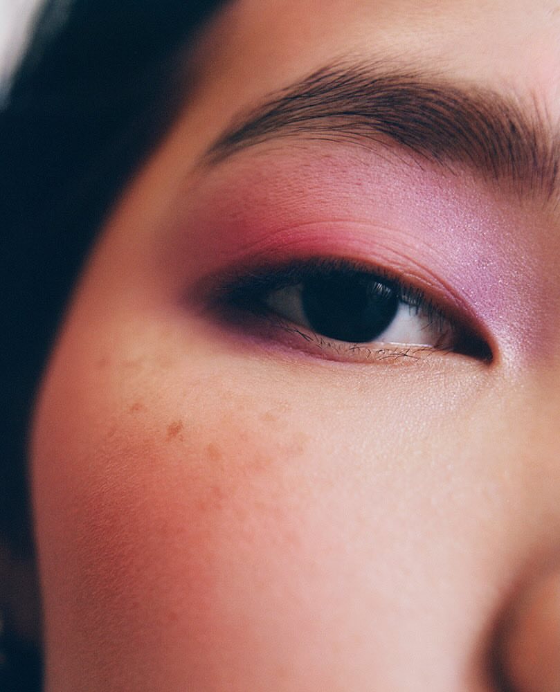 Close-up studio portrait of a woman's face features with violet and pink eyeshadow, defined eyebrow, and visible freckles on her cheek.